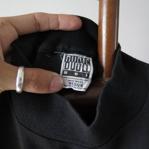 Op de afbeelding: Zwart T-shirt met een hoge hals en een wit label met de tekst "Bugle Boy" en "Medium" in zwarte tekst. Het label vermeldt ook "Made in USA" en "See reverse for care".