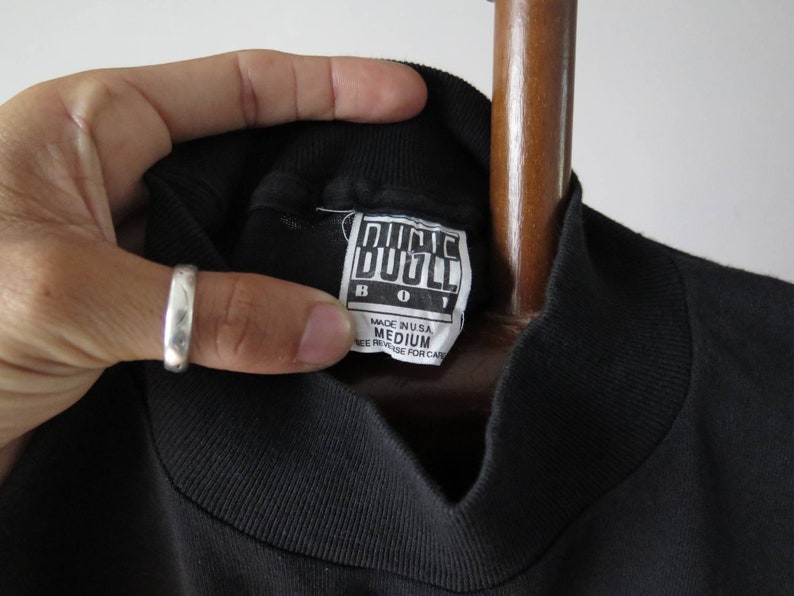 Op de afbeelding: Zwart T-shirt met een hoge hals en een wit label met de tekst "Bugle Boy" en "Medium" in zwarte tekst. Het label vermeldt ook "Made in USA" en "See reverse for care".