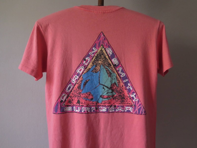 Pu&ograve; includere: Una maglietta color corallo con una grafica triangolare sul retro. La grafica include il testo "Gordon Smith Surfgear" e un design blu e verde acqua. La maglietta ha maniche corte e girocollo.