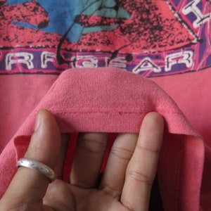 Pu&ograve; includere: T-shirt color corallo con un design grafico con la scritta "SURF GEAR" in lettere bianche. Il design include elementi rosa, viola e blu. Il colletto della maglietta &egrave; visibile.