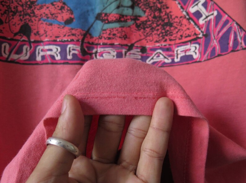 Pu&ograve; includere: T-shirt color corallo con un design grafico con la scritta "SURF GEAR" in lettere bianche. Il design include elementi rosa, viola e blu. Il colletto della maglietta &egrave; visibile.