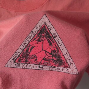 Pu&ograve; includere: Una maglietta color corallo con una grafica triangolare. La grafica presenta un design rosso e nero con le parole "Bordons Smith Surf Gear" sui bordi. La maglietta &egrave; realizzata in un materiale morbido, possibilmente di cotone.