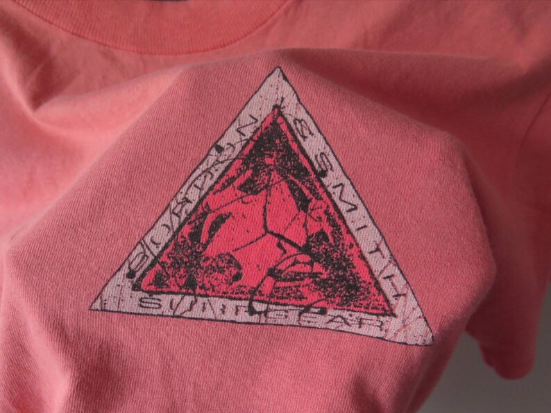 Pu&ograve; includere: Una maglietta color corallo con una grafica triangolare. La grafica presenta un design rosso e nero con le parole "Bordons Smith Surf Gear" sui bordi. La maglietta &egrave; realizzata in un materiale morbido, possibilmente di cotone.