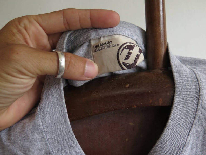 Pu&ograve; includere: Maglietta grigia con un'etichetta che recita "Dirt McGirt, Fashion Industry, Trademark Lifestyle Co."