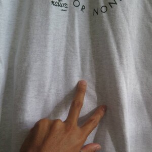 May include: A close-up of a gray t-shirt with the text "natura OR NON" printed on the front.