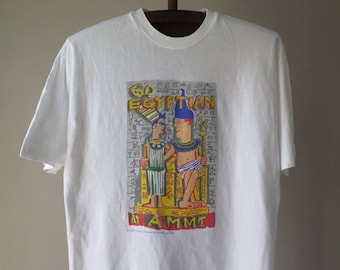 May include: White t-shirt with a colorful graphic print of two figures in Egyptian-style clothing. The text "Go Egyptian at AMM" is printed below the figures.