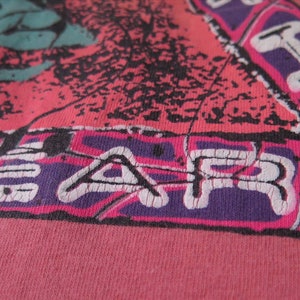 Pu&ograve; includere: Primo piano di una maglietta rosa con un design grafico. Il design presenta una combinazione di colori rosa, viola, nero e verde acqua. La parola "FEAR" &egrave; stampata in bianco, con uno stile vintage usurato.