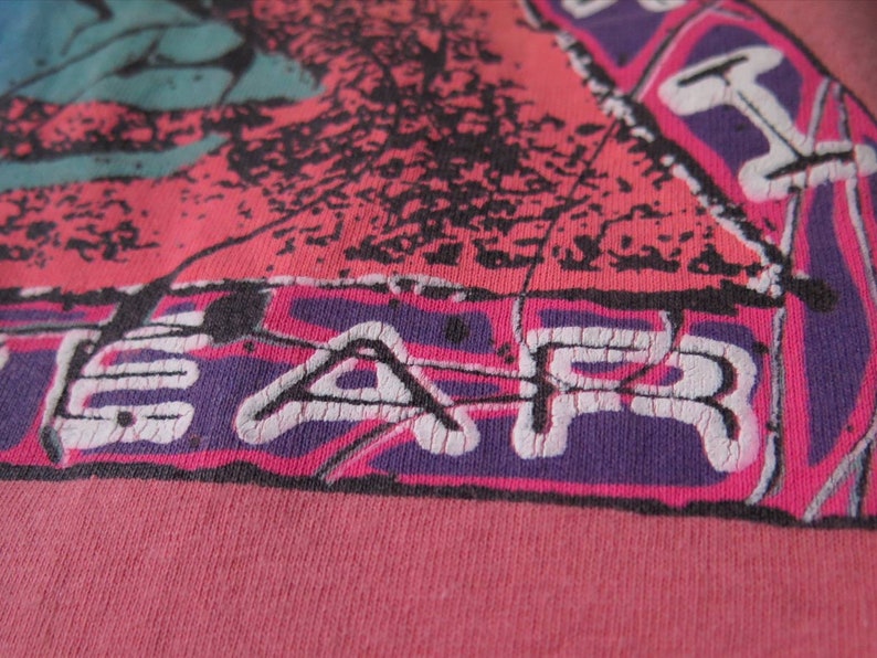 Pu&ograve; includere: Primo piano di una maglietta rosa con un design grafico. Il design presenta una combinazione di colori rosa, viola, nero e verde acqua. La parola "FEAR" &egrave; stampata in bianco, con uno stile vintage usurato.