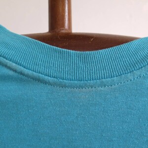 May include: Close-up of the neckline of a turquoise blue t-shirt. The neckline is ribbed and has a double-stitched seam.