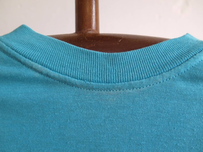May include: Close-up of the neckline of a turquoise blue t-shirt. The neckline is ribbed and has a double-stitched seam.