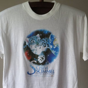 Puede incluir: Camiseta blanca con un gráfico azul y blanco de un cachorro de leopardo de las nieves en el espacio. El gráfico incluye un planeta azul y estrellas. El texto "Chim Schimmel" está impreso debajo del gráfico con las palabras "Love it put into the Mother Earth".