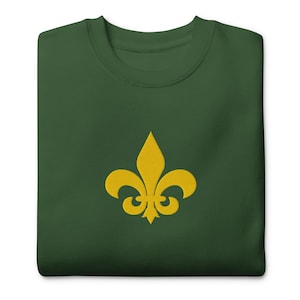 May include: A folded, dark green sweatshirt featuring a gold fleur-de-lis emblem embroidered on the front. The emblem is a stylized lily with three petals. The sweatshirt is made of a soft material.