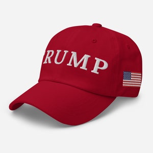 Puede incluir: Gorra de béisbol roja con la palabra "TRUMP" bordada en blanco en la parte delantera y un pequeño parche de la bandera estadounidense en el lateral.