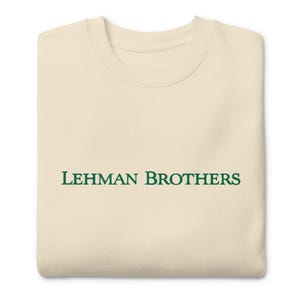May include: Cream-colored sweatshirt with the text "LEHMAN BROTHERS" in green, centered on the front. The crew neck sweatshirt is folded neatly, showcasing the design. The garment appears to be made of a soft, comfortable material.