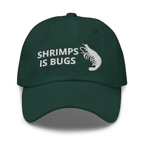 Puede incluir: Una gorra de béisbol verde oscuro con bordado blanco que dice "SHRIMPS IS BUGS" y un diseño de camarón bordado en blanco.