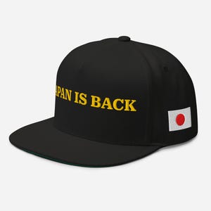 以下が含まれることがあります： 黒いベースボールキャップ。つばは平らで、黄色で「JAPAN IS BACK」と刺繍されています。側面に日本の国旗のパッチが付いています。つばの裏側は緑色です。