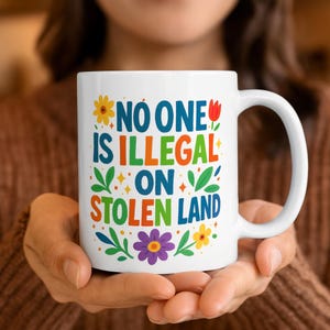 May include: White ceramic mug with the text "NO ONE IS ILLEGAL ON STOLEN LAND" in colorful letters and floral accents. The mug is held by a person. The mug is a standard size for coffee or tea.
