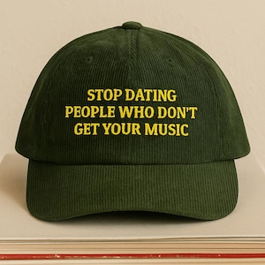 May include: A dark green corduroy baseball cap with the embroidered text "STOP DATING PEOPLE WHO DON'T GET YOUR MUSIC" in yellow. The cap is sitting on top of a stack of books.