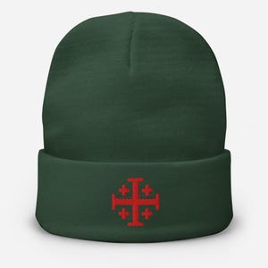 Könnte beinhalten: Dunkelgrüne Beanie-Mütze mit einem roten Jerusalemer Kreuz auf der Vorderseite. Die Mütze ist aus weichem Strickmaterial gefertigt und hat einen umgeschlagenen Saum. Das Jerusalemer Kreuz ist ein christliches Symbol.