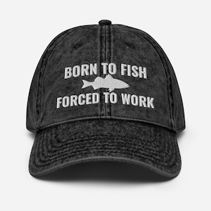 以下が含まれることがあります： 白い刺繍で「Born to Fish Forced to Work」と書いてあり、白い魚のシルエットが描かれた、黒のダメージ加工デニムのベースボールキャップです。