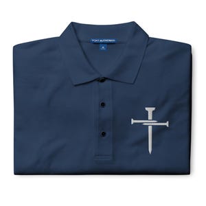 May include: Navy blue polo shirt with a white embroidered cross and nail design. The shirt has a collared neckline, three buttons, and the "Port Authority" brand label. A medium size is indicated on the label.