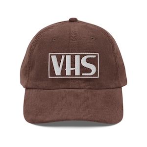 May include: A brown corduroy baseball cap featuring the white embroidered letters "VHS". The cap has a curved peak and an adjustable strap. This retro-style hat is a nod to the past.