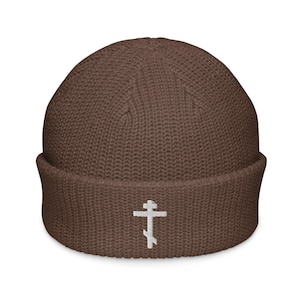 May include: A brown knit beanie with a folded brim. The beanie features a white embroidered Eastern Orthodox cross on the front. The hat is made of a ribbed knit material.