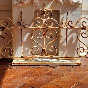 Puede incluir: Una verja decorativa de metal de estilo antiguo con un acabado blanquecino desgastado. La verja presenta adornos de volutas y diseños en forma de corazón. Se encuentra sobre un fondo de pared blanca con molduras decorativas y suelo de madera.