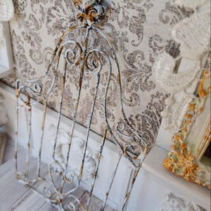 Puede incluir: Una puerta decorativa de metal blanco desgastado con adornos de volutas y un acento de flor de lis. La puerta tiene una apariencia oxidada y envejecida, lo que sugiere un encanto vintage o antiguo. La puerta está diseñada para fines decorativos.