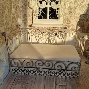 Puede incluir: Una cama de metal en miniatura y ornamentada con un cojín color crema. El marco de la cama presenta volutas y un ribete de encaje decorativo en la parte inferior. La escena está ambientada en una casa de muñecas con papel pintado y una ventana.