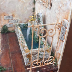 May include: Miniature diorama of a stone fountain with a golden faucet and flowing water. The fountain is set against a textured, off-white wall with a small window. A decorative, wrought-iron railing frames the fountain.