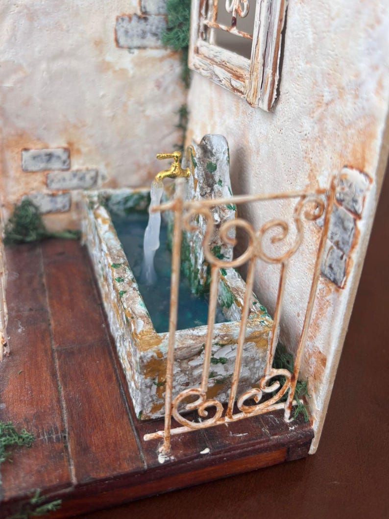 May include: Miniature diorama of a stone fountain with a golden faucet and flowing water. The fountain is set against a textured, off-white wall with a small window. A decorative, wrought-iron railing frames the fountain.