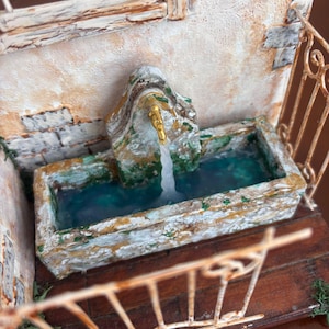 May include: A miniature fountain with a stone-like appearance, featuring a golden faucet and water flowing into a rectangular basin. The fountain is set against a textured wall with a window and a decorative railing.