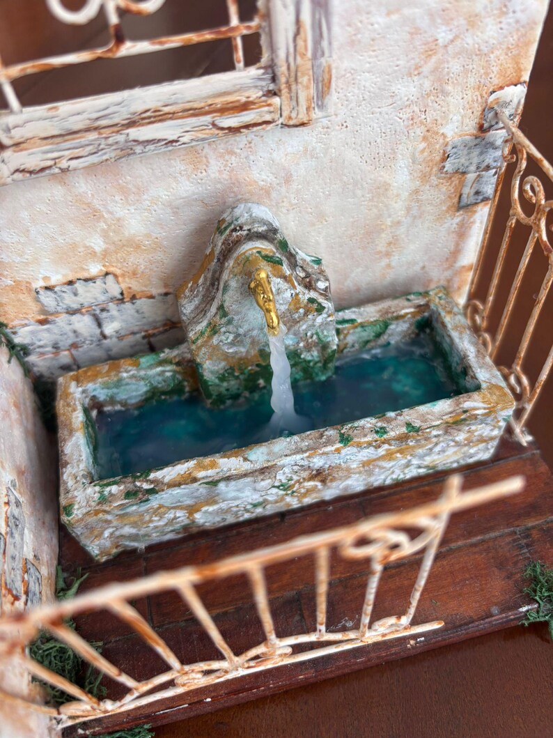 May include: A miniature fountain with a stone-like appearance, featuring a golden faucet and water flowing into a rectangular basin. The fountain is set against a textured wall with a window and a decorative railing.