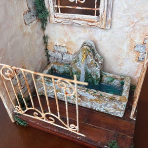 May include: Miniature diorama of a rustic stone fountain with a small balcony. The fountain has a weathered appearance with a water feature. The balcony has a decorative wrought iron railing. The scene is set against a textured wall with a window.