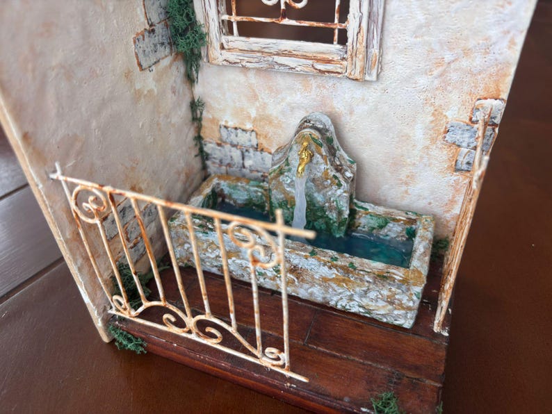 May include: Miniature diorama of a rustic stone fountain with a small balcony. The fountain has a weathered appearance with a water feature. The balcony has a decorative wrought iron railing. The scene is set against a textured wall with a window.