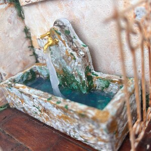 May include: A miniature fountain with a weathered, stone-like appearance. The fountain features a basin filled with blue water, a textured spout, and golden faucets evoking a rustic aesthetic.