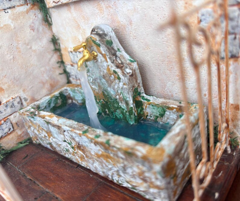 May include: A miniature fountain with a weathered, stone-like appearance. The fountain features a basin filled with blue water, a textured spout, and golden faucets evoking a rustic aesthetic.