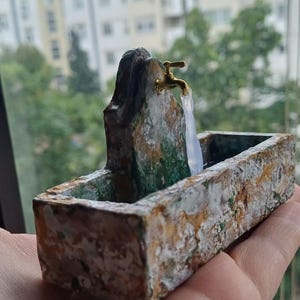 May include: A miniature fountain with a gold-colored faucet and a rectangular basin. The fountain is made of stone-like material with green and white accents. The water appears to be flowing from the faucet.