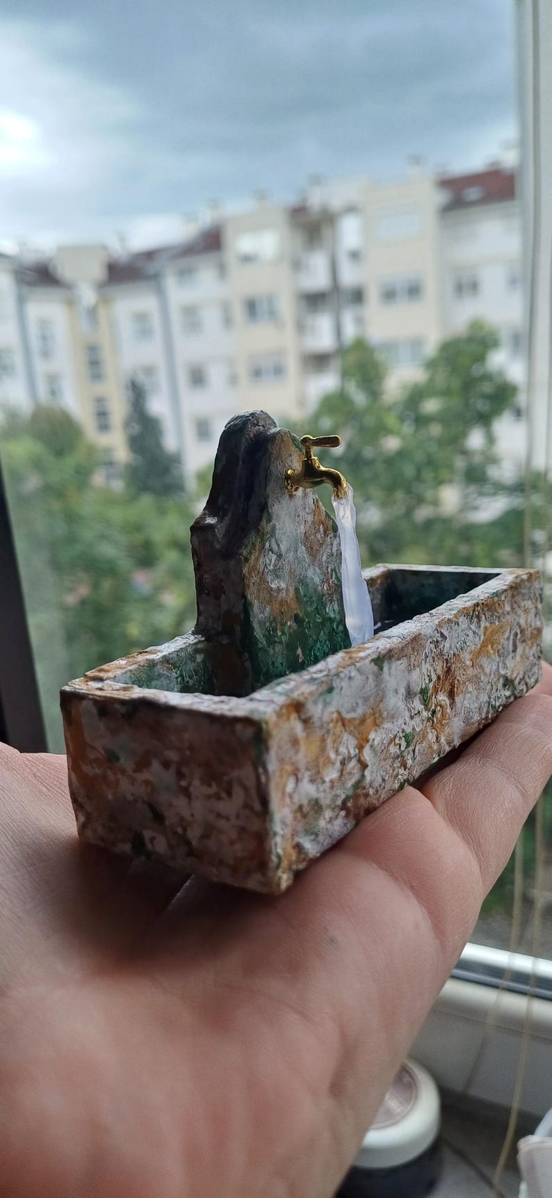 May include: A miniature fountain with a gold-colored faucet and a rectangular basin. The fountain is made of stone-like material with green and white accents. The water appears to be flowing from the faucet.