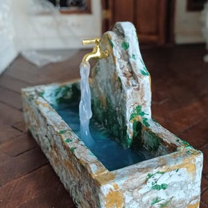 May include: Miniature stone fountain with a gold faucet and flowing water. The rectangular basin is painted with white, yellow, and green accents. The fountain is set on a dark wood floor, creating a charming, rustic aesthetic.