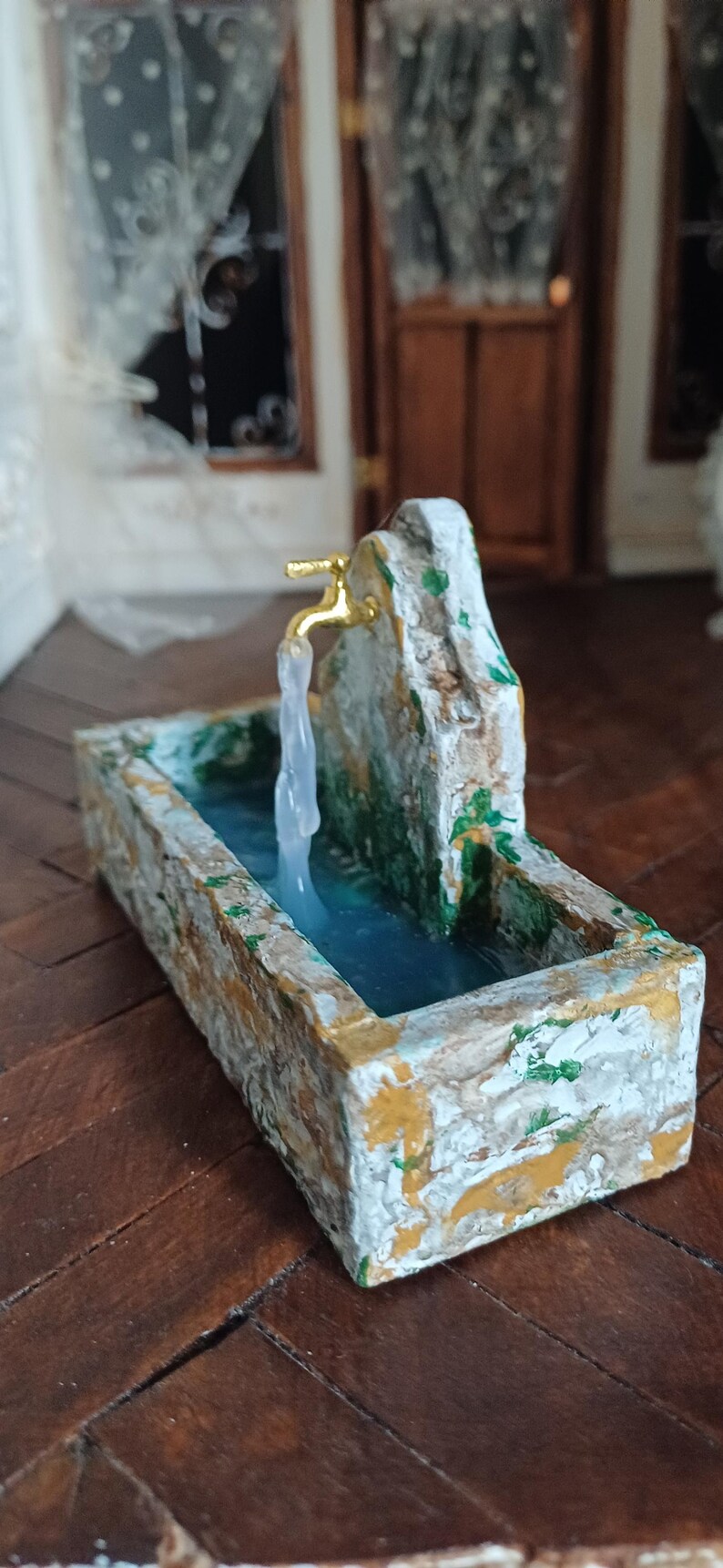 May include: Miniature stone fountain with a gold faucet and flowing water. The rectangular basin is painted with white, yellow, and green accents. The fountain is set on a dark wood floor, creating a charming, rustic aesthetic.