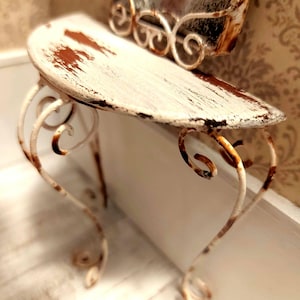 May include: A small, distressed white metal table with a curved tabletop and ornate, scrolled legs. The tabletop has areas of exposed brown, adding to the vintage aesthetic. The table is likely a decorative accent piece.