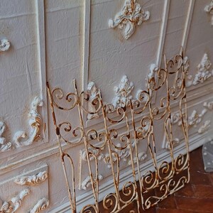May include: Two cream-coloured wrought iron gates with a distressed finish, leaning against a textured white wall with decorative moulding. The gates feature scrolled designs and a rusty patina, suggesting an antique or vintage aesthetic.