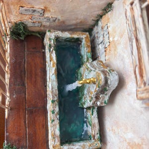 May include: Miniature fountain with a rectangular basin filled with teal water. The fountain has a stone-like appearance with gold accents and a decorative spout. The surrounding walls and floor are textured, with wooden planks and brick details.