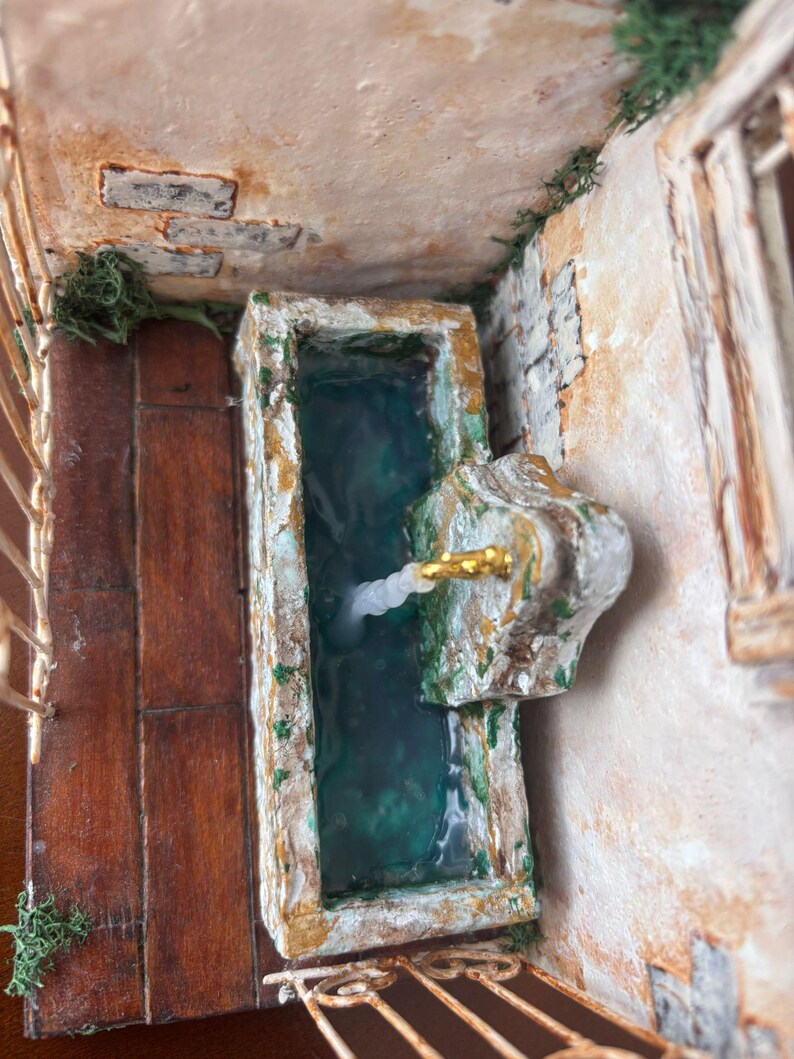 May include: Miniature fountain with a rectangular basin filled with teal water. The fountain has a stone-like appearance with gold accents and a decorative spout. The surrounding walls and floor are textured, with wooden planks and brick details.