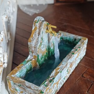 May include: Miniature fountain with a gold faucet and flowing water. The fountain is made of stone-like material with green and gold accents. The water flows into a rectangular basin, creating a serene, decorative piece.