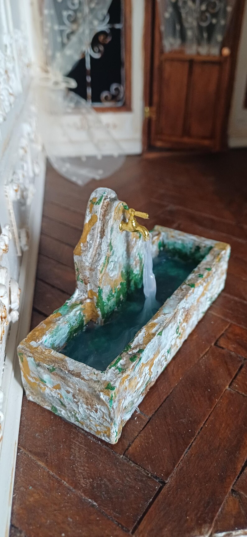 May include: Miniature fountain with a gold faucet and flowing water. The fountain is made of stone-like material with green and gold accents. The water flows into a rectangular basin, creating a serene, decorative piece.