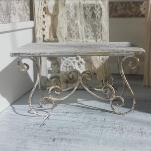 May include: A miniature decorative table in white and gold, featuring a rectangular top and scrolled legs. The table has a distressed, vintage look, with a weathered white finish and gold accents. It is designed for decorative use, possibly for a dollhouse.