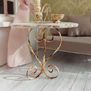 Pode incluir: Uma mesa em miniatura, branca e desgastada, com uma base de ferro forjado em forma de coração. A mesa tem um tampo redondo e é adornada com uma pequena tigela decorativa e um castiçal. A cena é ambientada numa casa de bonecas.